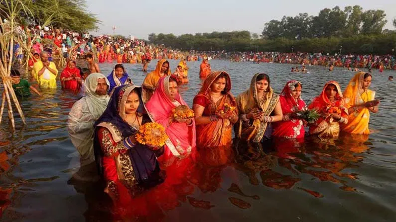 Hindu women offering Arghya to rising Sun during Chhath Puja, standing in river with traditional saree and bamboo baskets filled with fruits and diyas.