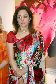 Hema Malini ka close-up shot, red aur multi-colored floral print ki saree mein, ek event mein khadi hain.